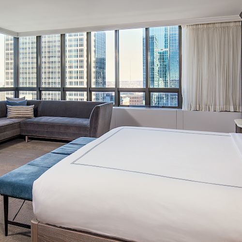 A modern, well-lit hotel room with a large bed, a dark blue sofa, a small side table, and large windows offering a cityscape view, ending the sentence.