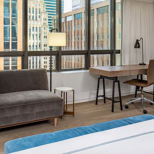 A modern hotel room with a view of city buildings, featuring a sofa, floor lamp, desk with a chair, and part of a bed with blue bedding.