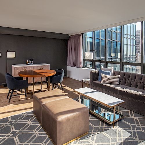 A modern living room features large windows, a gray couch, a rectangular coffee table, a dining area with a round table, and various wall art.