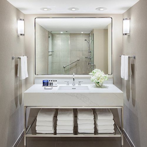 A modern bathroom with a large mirror, sink, neatly folded towels beneath, wall-mounted lights and a shower seen in the reflection.