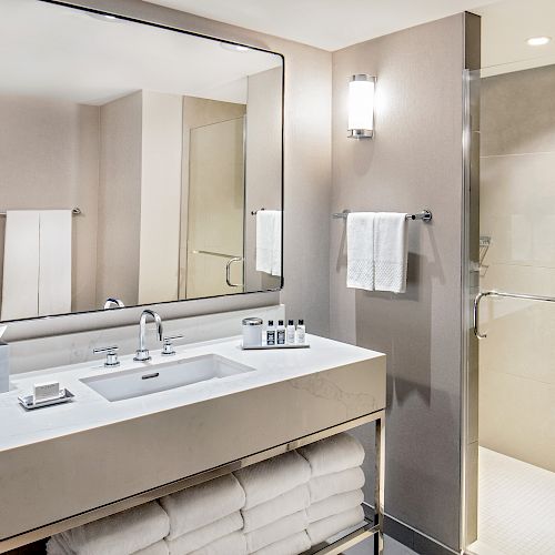 A modern bathroom with a large mirror, sink, toiletries, towel racks, and a walk-in shower. White towels and folded towels are stored below.