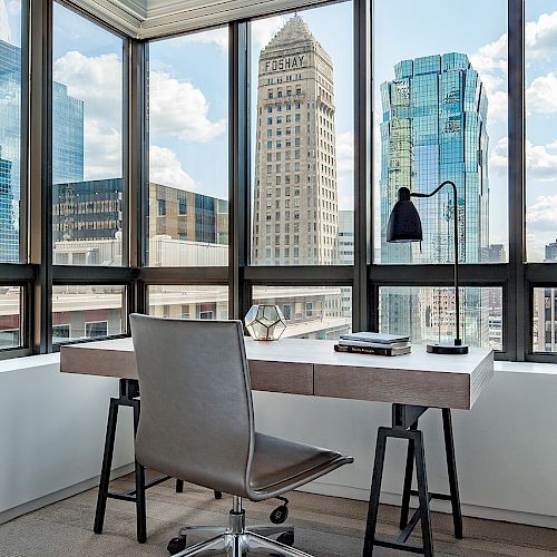 A modern office setup with a desk, chair, and lamp by large windows overlooking a cityscape with tall buildings under a partly cloudy sky.