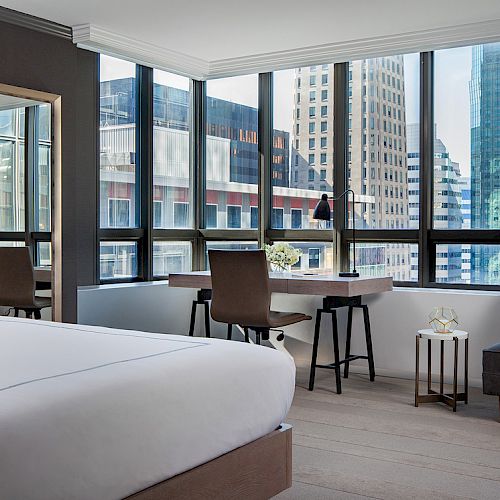 A modern hotel room with a large bed, seating area, desk, and floor-to-ceiling windows offering a city view with tall buildings outside.