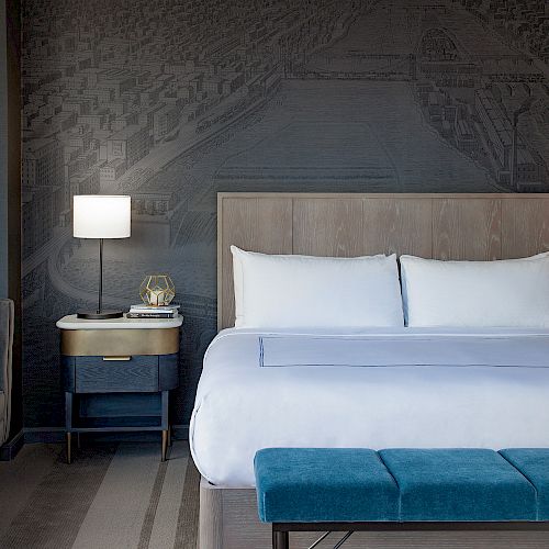 A modern hotel room features a neatly made bed, nightstands with lamps, a gray sofa, and a blue bench at the foot of the bed.