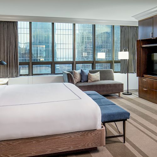 A modern hotel room with a large bed, blue bench, sofa, flat-screen TV, and large windows with city views, featuring contemporary decor.