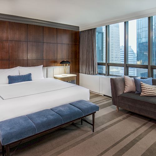 A modern hotel room features a bed, cushioned bench, sofa, and large windows with a city view, with warm wood paneling and contemporary decor.