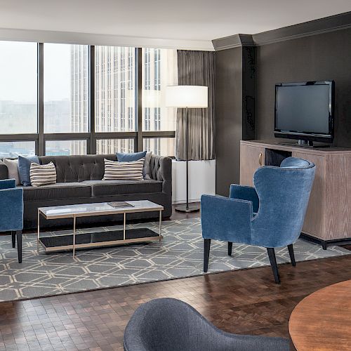 A modern living room with blue chairs, a gray couch, a TV on a stand, two floor lamps, and a patterned rug by large windows with city views.