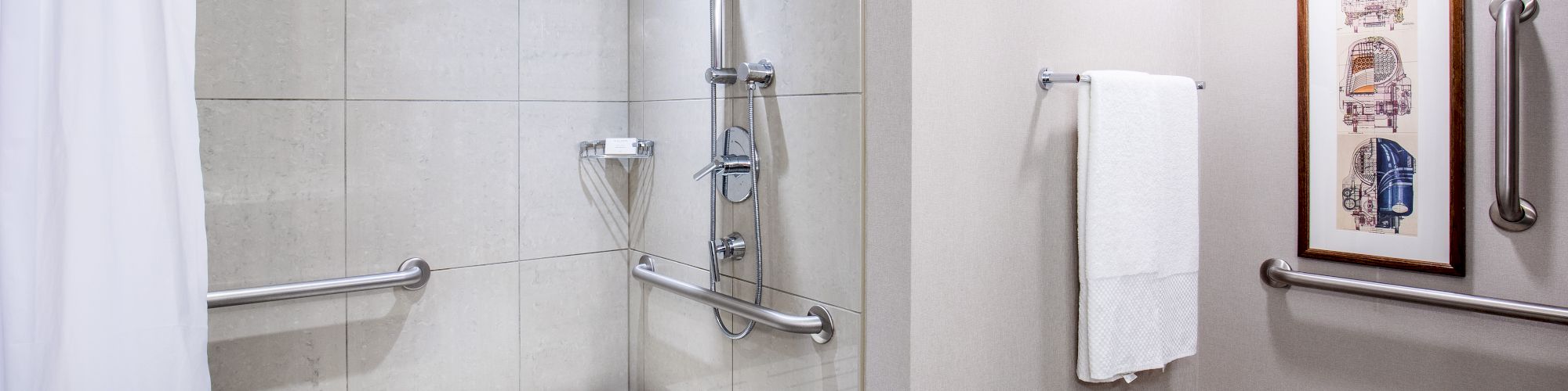 This image shows an accessible bathroom with a shower area including a bench, grab bars, a showerhead, a towel, and a framed picture on the wall.