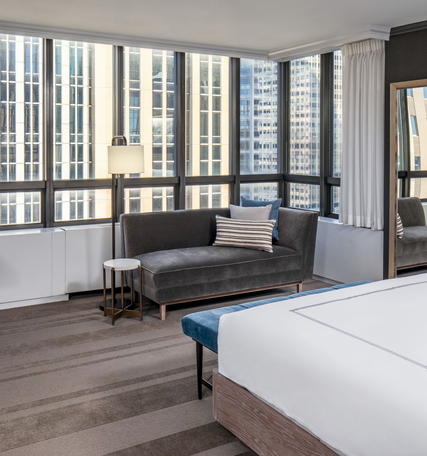 The image shows a modern hotel room with a large bed, a sofa, a desk, and a floor-to-ceiling window with a cityscape view.