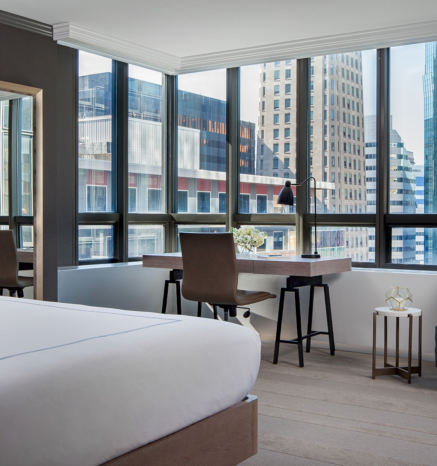 A modern hotel room features a bed, desk, and seating area with large windows overlooking skyscrapers and urban scenery.