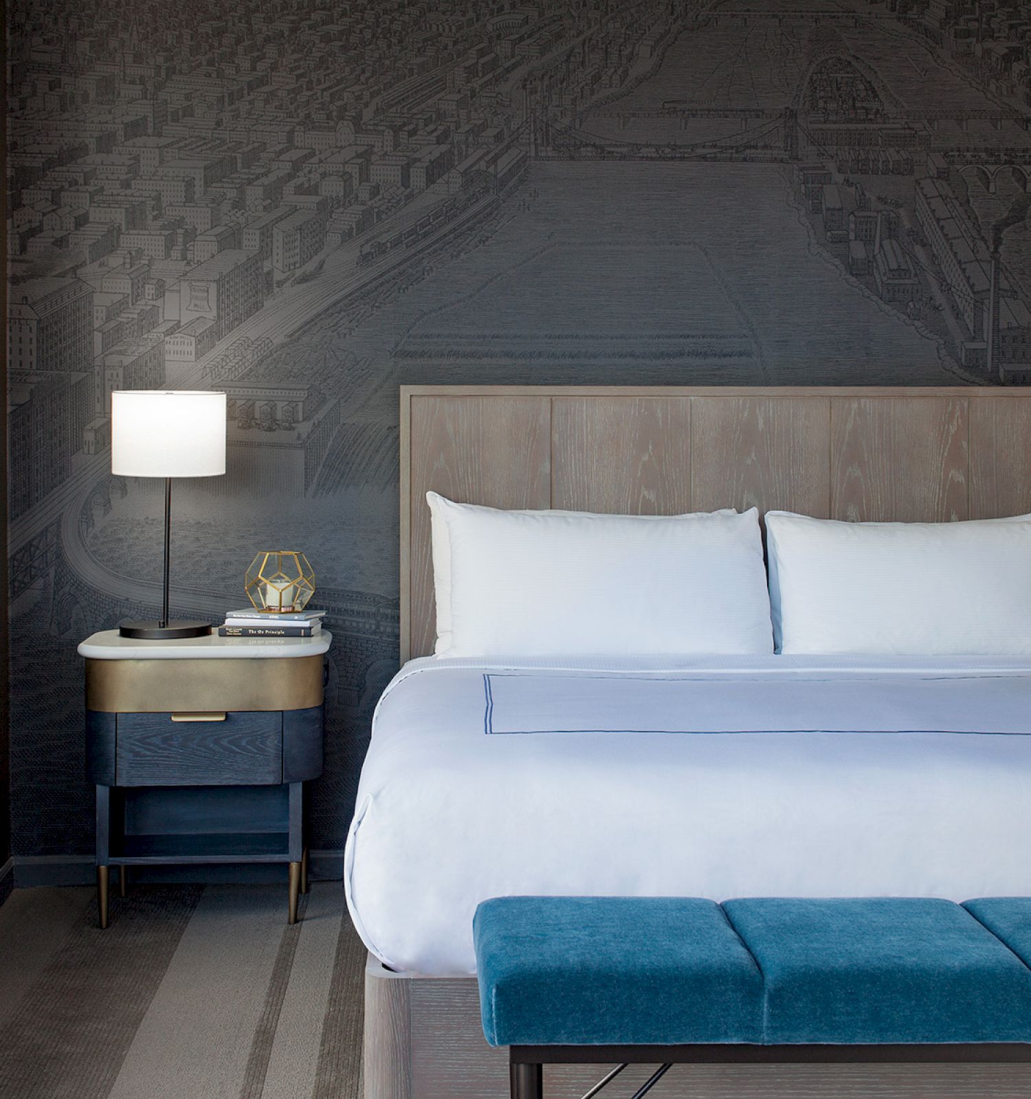 A modern hotel room features a king-sized bed, blue bench, nightstands with lamps, and a sofa, with a large mural on the back wall ending the sentence.